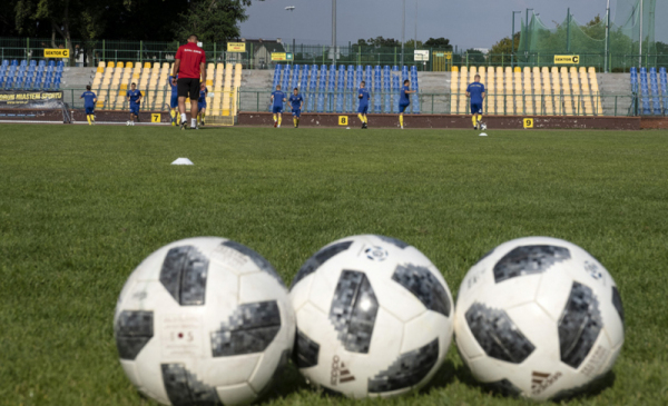 Na zdjęciu widać trzy piłki do gry w piłkę nożną, leżące na murawie, w tle widać trybuny polamowane na żółto-niebiesko.