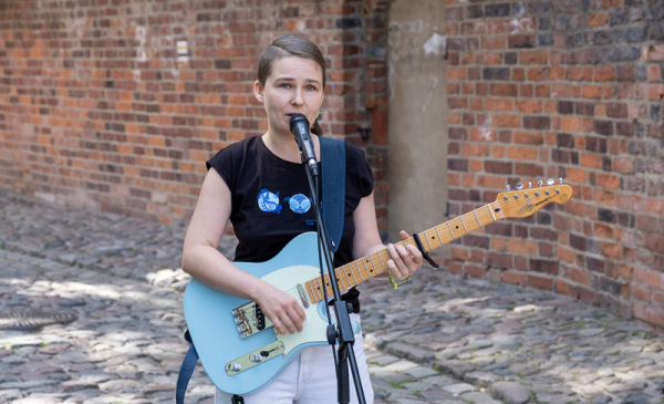 Na zdjęciu: młoda dziewczyna śpiewa do mikrofony, gra na niebieskiej gitarze elektrycznej, w tle ceglany mur