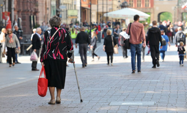 Na zdjęciu widać starszą panią, poruszającą się o kuli, niosącą zakupy w czerwonej torbie