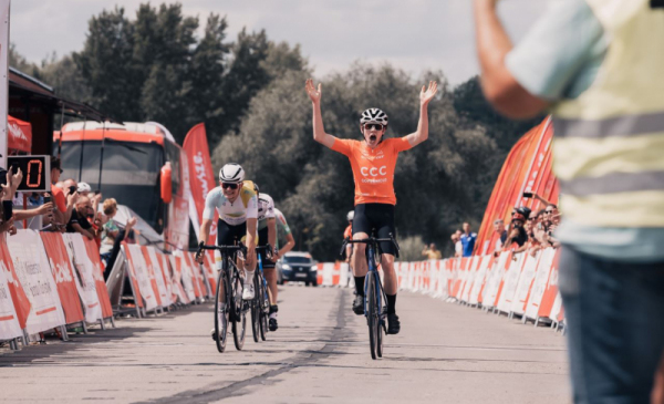 Na zdjęciu: zawodnik kolarski w pomarańczowej koszulce wjeżdża na metę i unosi ręce w geście zwycięstwa
