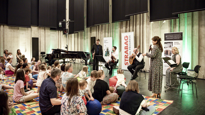 grupa dzieci i rodziców siedzi na kolorowym dywanie w sali prób orkiestry, przed nimi muzycy, kobieta z mikrofonem prowadzi koncert i śmieje się