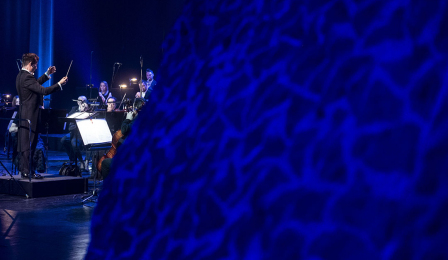 Dyrygent Adam Banaszak dyryguje orkiestrą, na pierwszym planie fragment charakterystycznej ściany w CKK Jordanki