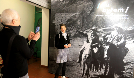 Mężczyzna robi zdjęcie kobiecie przy dużej fotografii czarnobiałej, przedstawiającej podróżników na koniach