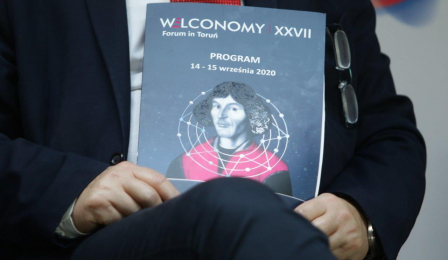 Uczestnik panelu dyskusyjnego trzyma na kolanach teczkę z napisem Welconomy Forum i podobizną Mikołaja Kopernika